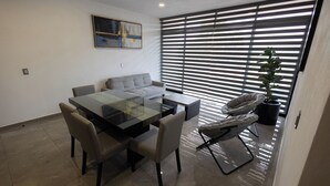 Un espace repas moderne avec une table en verre, des chaises rembourrées, un canapé et une grande fenêtre équipée de stores.