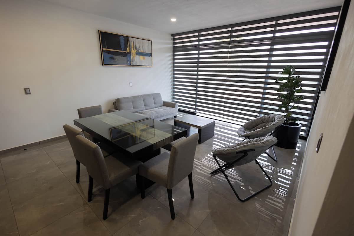 Un comedor moderno con una mesa de vidrio, sillas acolchadas, un sofá y un amplio ventanal con persianas.