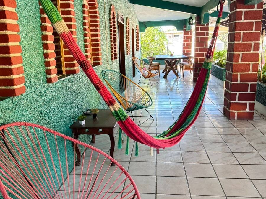 A covered patio area with a tiled floor, brick walls, and a hammock. There are metal chairs and a table in the background.