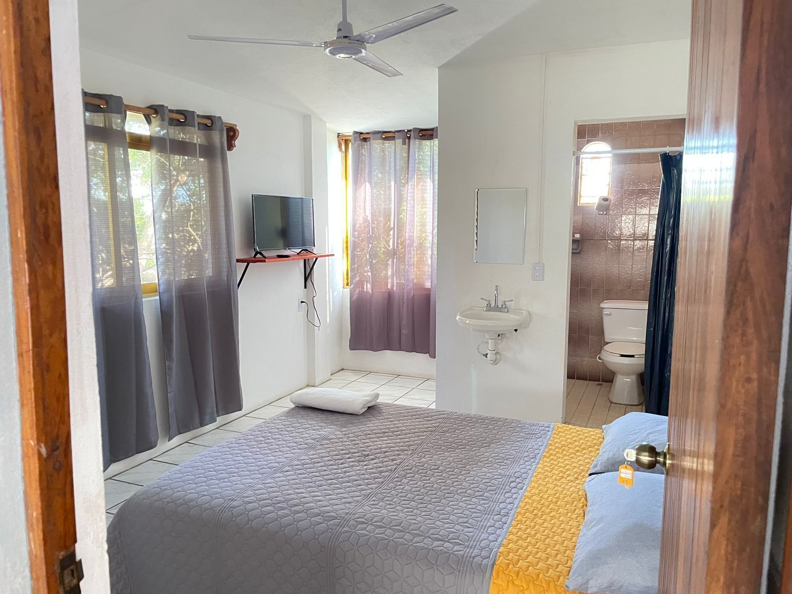 Een slaapkamer met een bed, een televisie op een dressoir, een nachtkastje, een raam met gordijnen, een badkamer met wastafel en toilet, en een plafondventilator.