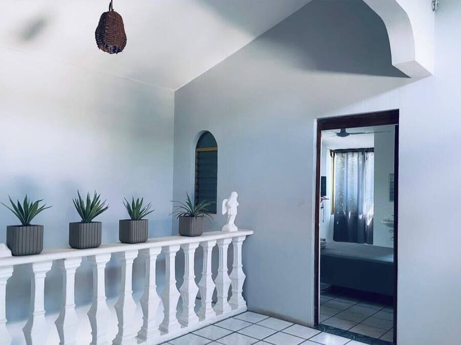 Un pasillo con barandilla blanca, plantas en macetas, una lámpara colgante y un vano que da a una habitación con cama y cortinas.