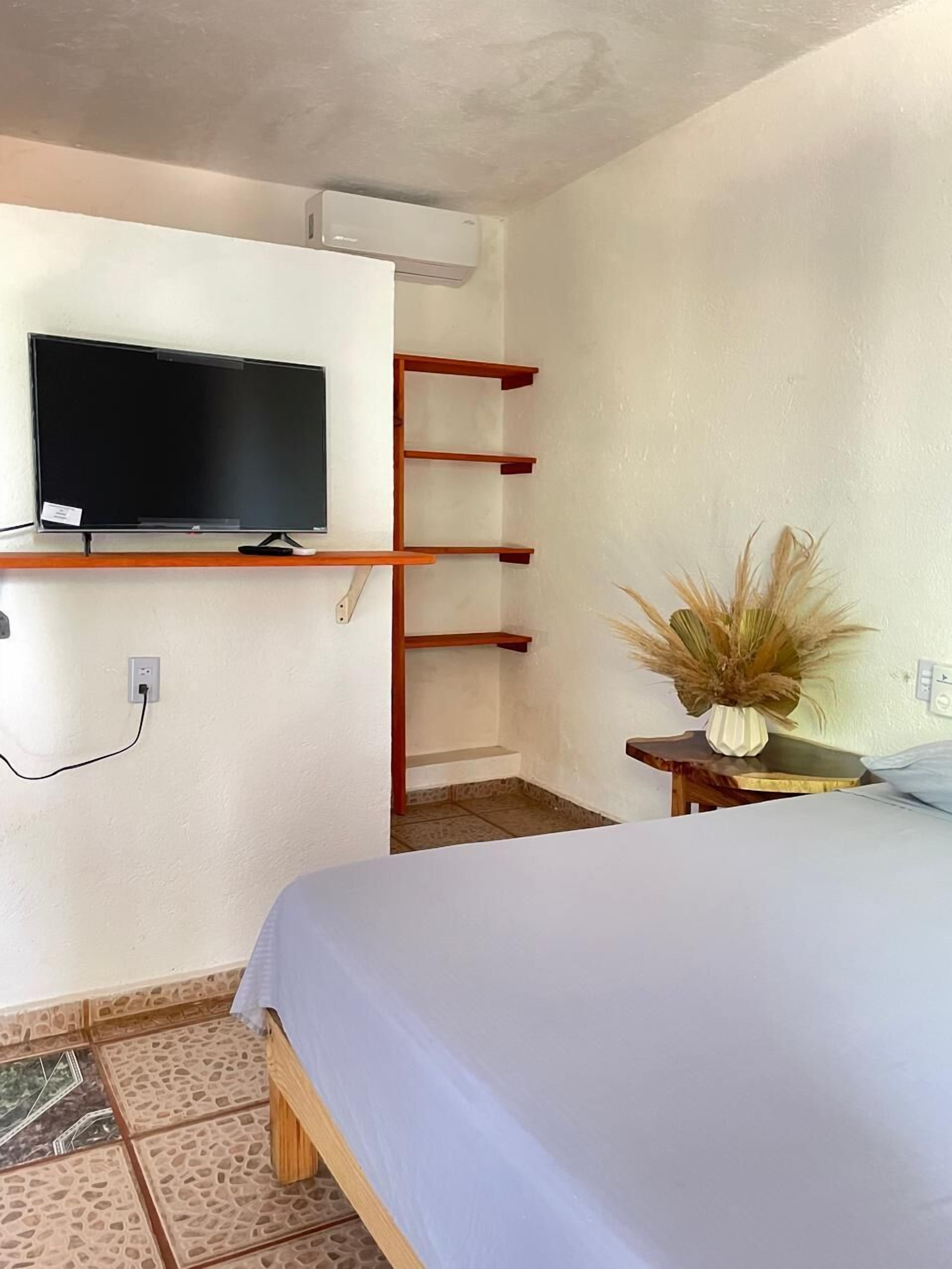 Un dormitorio con cama, televisor montado en la pared, estantería de madera y una mesita con un jarrón de plantas secas.