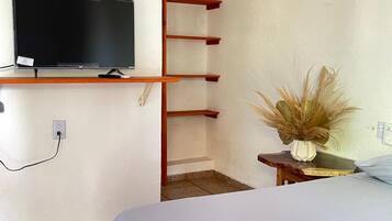 Un dormitorio con cama, televisor montado en la pared, estantería de madera y una mesita con un jarrón de plantas secas.
