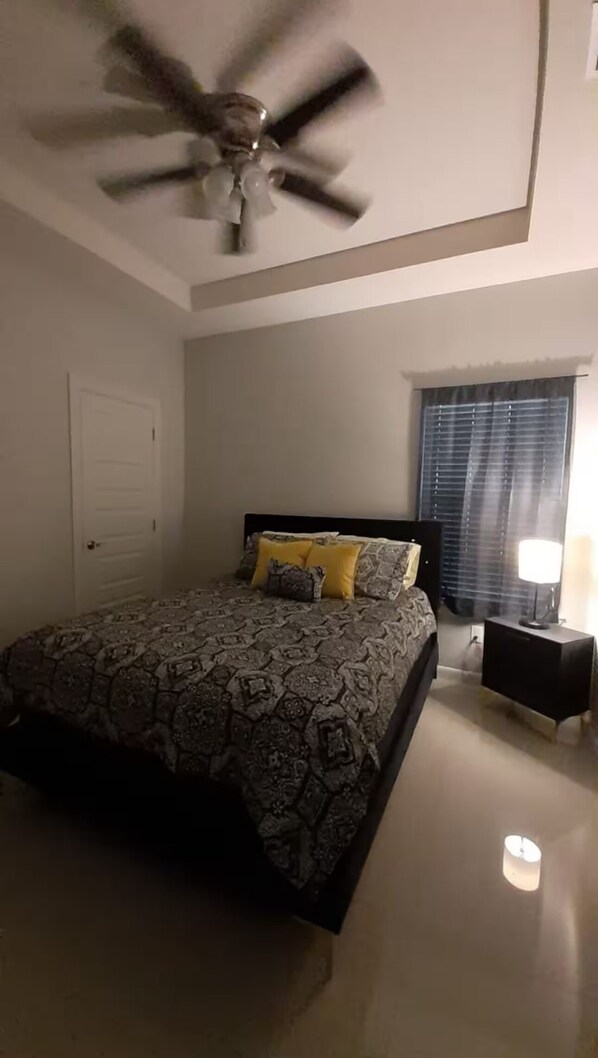 Ein Schlafzimmer mit einem Bett, einem Deckenventilator, einem Fenster mit Jalousien und einem Nachttisch mit einer Lampe.