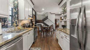 Una cocina moderna con muebles blancos, electrodomésticos de acero inoxidable, una mesa de comedor de madera y una escalera que sube a un nivel superior.