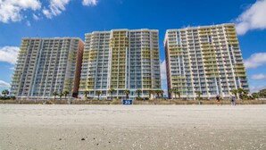 Un hotel de gran altura con varios pisos, una playa arenosa al frente y un cielo azul despejado.