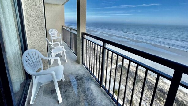 Un balcón con sillas de plástico blanco, piso de concreto y vista a la playa.