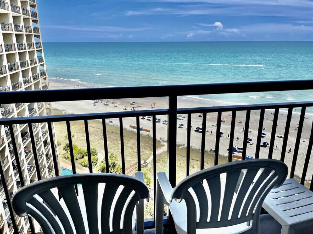 A balcony with two chairs overlooking a beach with a view of the ocean and a parking lot.