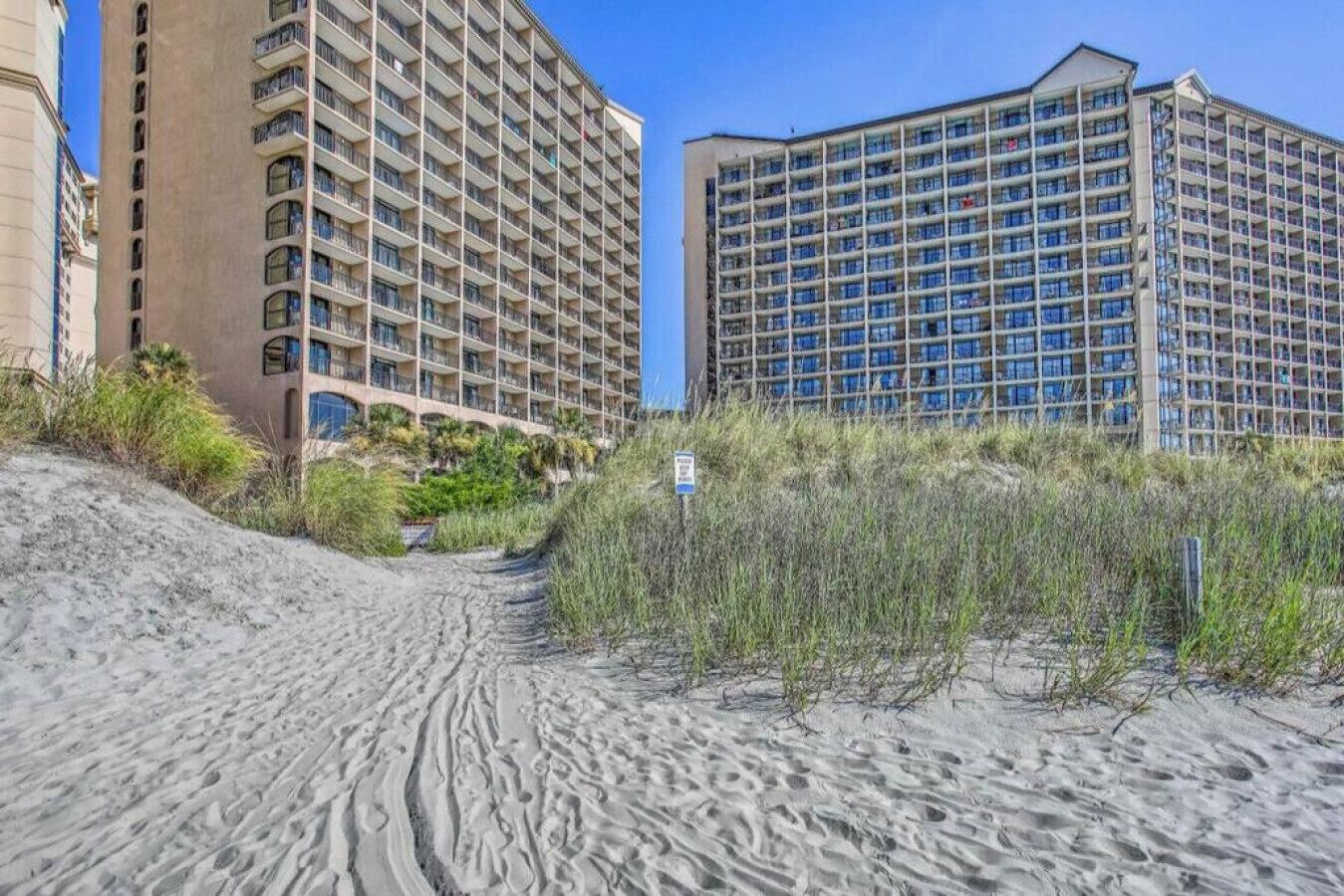 A beachfront hotel with multiple floors and balconies, surrounded by sandy terrain and sparse vegetation.