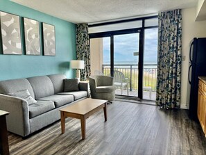 A living room with a sofa, armchair, and coffee table, a balcony with curtains, and a view of the outdoors. - Tropical Resort, Beachfront Condo with amazing oceanfront views!! 322 (North Myrtle Beach)