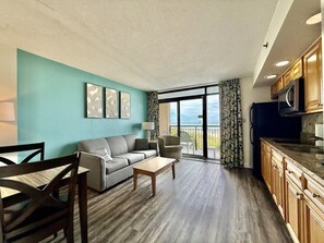 A modern living room with a sofa, a coffee table, and a dining area with chairs. There is a kitchen with wooden cabinets and a microwave. The room has a large window with a view of the outdoors. - Tropical Resort, Beachfront Condo with amazing oceanfront views!! 322 (North Myrtle Beach)