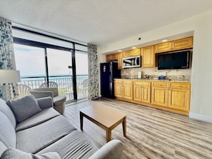 A modern living room with a sofa and a coffee table, a kitchen with wooden cabinets and a flat-screen TV, and a balcony with a view of the beach. - Tropical Resort, Beachfront Condo with amazing oceanfront views!! 322 (North Myrtle Beach)