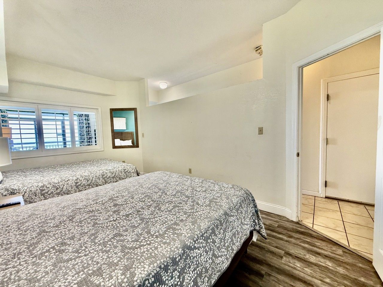 A bedroom with two beds, a window with blinds, a door, and a wooden floor.
