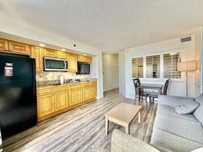 A modern kitchen with wooden cabinets, a black refrigerator, and a microwave. There is a dining area with a table and chairs, and a gray sofa.