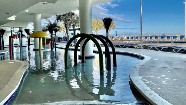 An indoor pool area with clear blue sky visible through large windows, surrounded by palm trees and a curved pool edge. - Right next to family Kingdom! Oceanfront 1br full kitchen washer/dryer 1107 (Myrtle Beach)
