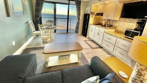 A modern living room with a sofa, a coffee table, and a dining area with a table and chairs. There is a kitchen area with white cabinets and a microwave. The room has a large window with a view of the ocean. - Tropical Resort, Modern Beachfront Condo with amazing oceanfront views!! 1018 (North Myrtle Beach)