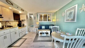 A compact kitchen with white cabinets, a microwave, and a coffee maker. There is a dining area with a table and chairs, and a window with blinds. - Tropical Resort, Modern Beachfront Condo with amazing oceanfront views!! 1018 (North Myrtle Beach)