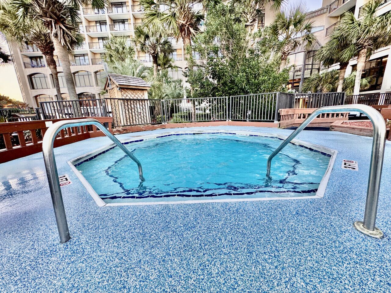 A swimming pool with a blue tiled floor, surrounded by a metal fence, and palm trees in the background.