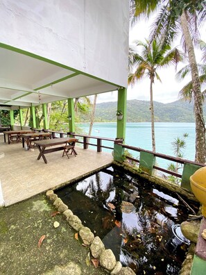 A covered outdoor seating area with tables and chairs, a small pond with rocks, and a view of a lake and mountains.