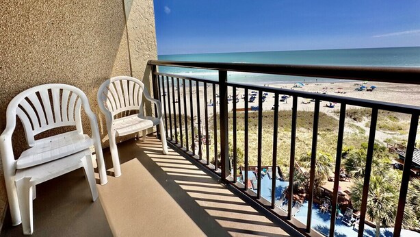 A balcony with two white plastic chairs, a pool, and a beach view. - Tropical Resort, Beachfront Condo with amazing oceanfront views!! 609 (North Myrtle Beach)