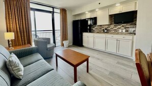 Uma sala de estar com sofá, mesa pequena e cozinha equipada, incluindo armários e televisão.