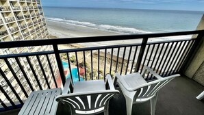 Varanda com duas cadeiras de plástico brancas e uma mesa, com vista para uma praia e oceano visível, e um edifício ao fundo.