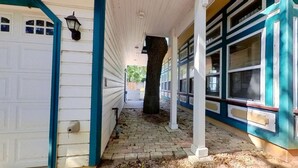 A covered porch with a white garage door, a tree trunk in the center, and a blue and white building with multiple windows. - The Buttercup Cottage (San Marcos)