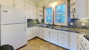 A kitchen with white cabinets, a granite countertop, a double sink, and a window with a view of trees. - The Buttercup Cottage (San Marcos)