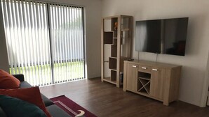 A living room with a sofa, a TV, a wooden cabinet, and a large window with blinds.
