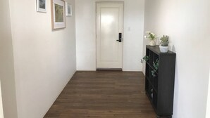 A hallway with a wooden floor, white walls, and a door at the end. There are framed pictures on the wall and a plant on a shelf.