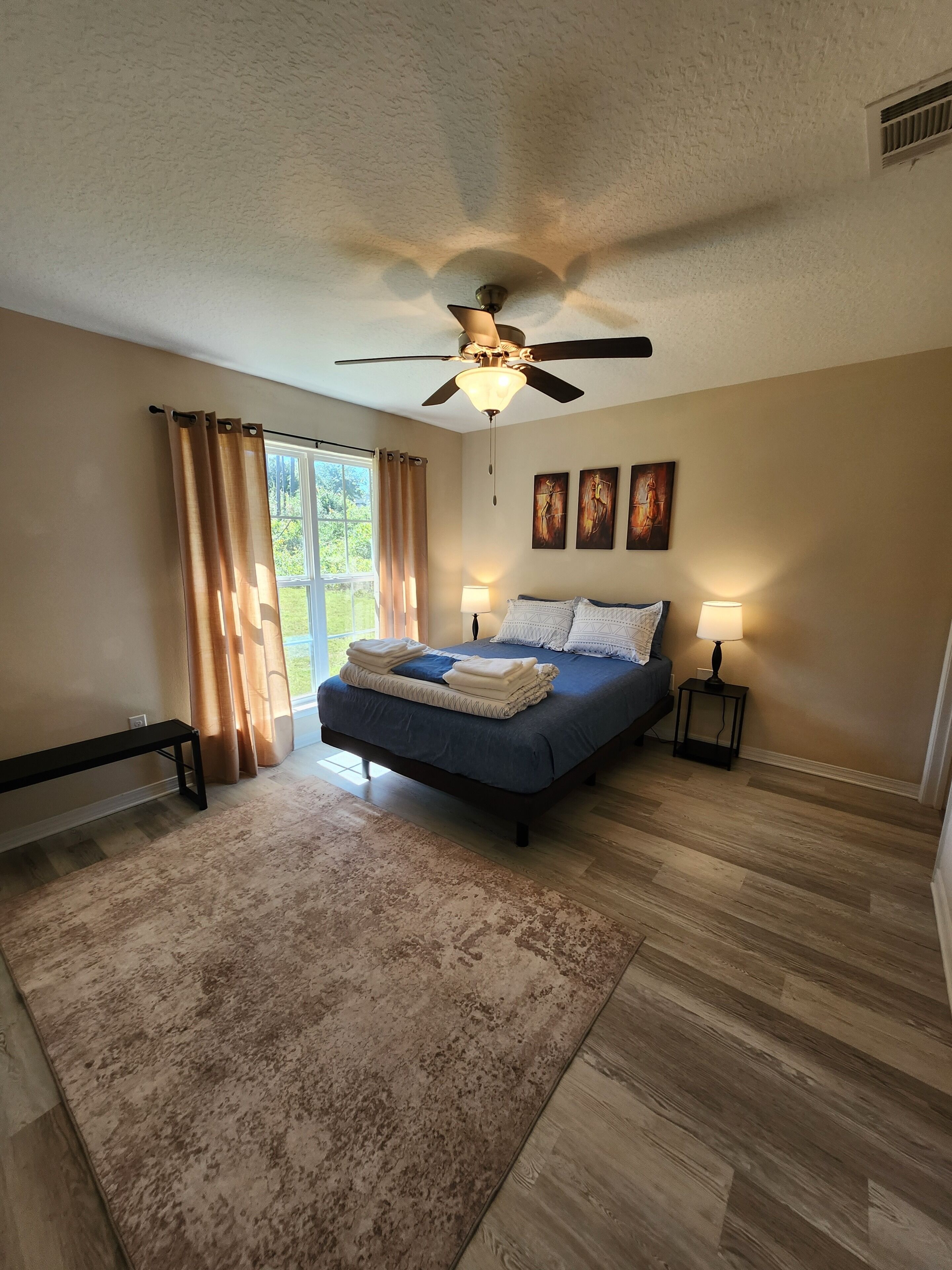 Une chambre à coucher avec un lit, un ventilateur de plafond, un banc, une fenêtre avec des rideaux et un tapis.