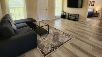 Un salon spacieux avec un canapé d’angle aux couleurs foncées, une table basse en verre, un tapis à motifs et un ventilateur de plafond.