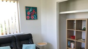 A living room with a black leather sofa, two blue cushions, a small round table, and a wooden bookshelf. - Marina Retreat Geraldton - Centrally Located in Garden Setting (Beresford)