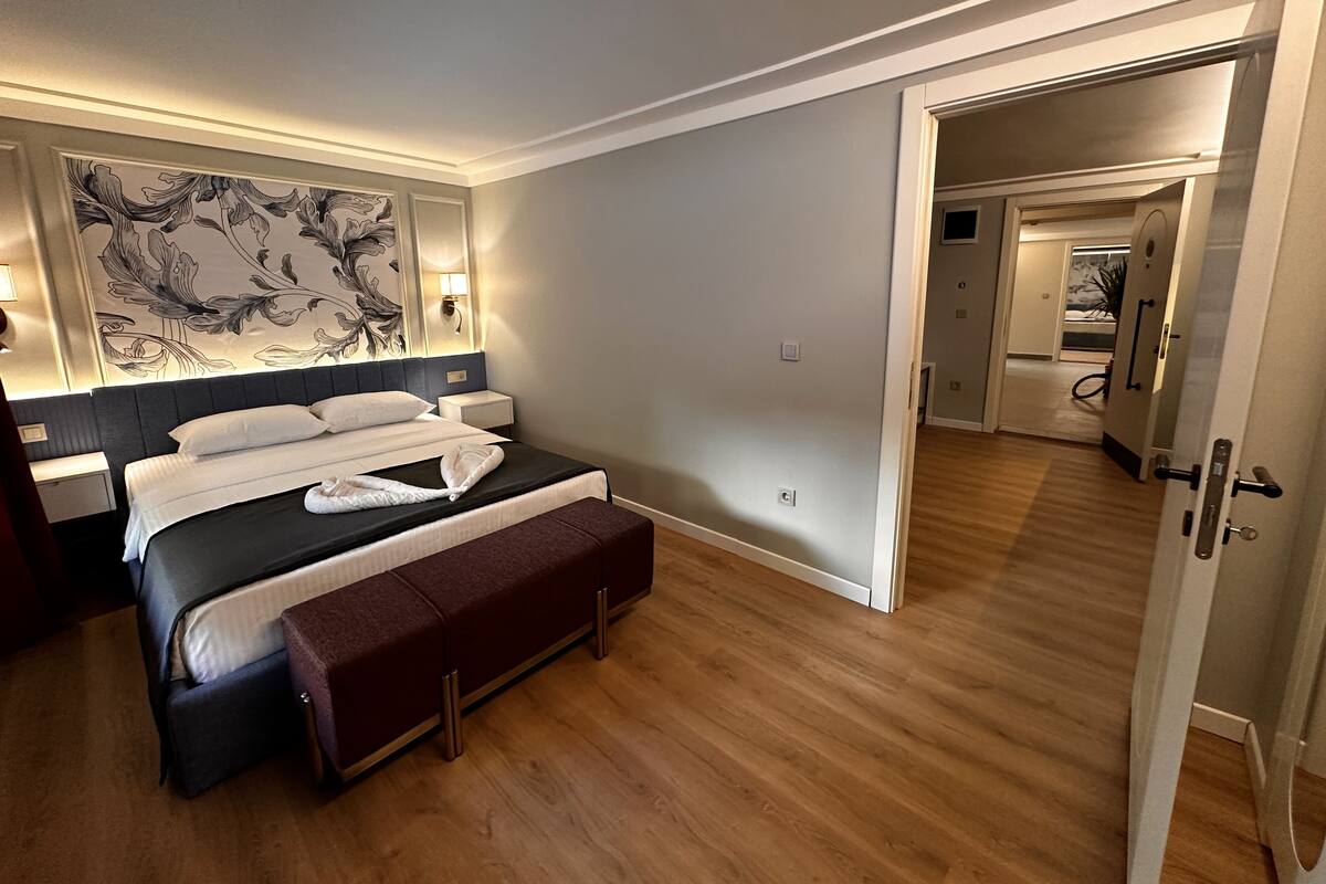 A modern hotel room with a large bed, bedside tables, a bench, a decorative wall, and a view into the adjacent room.