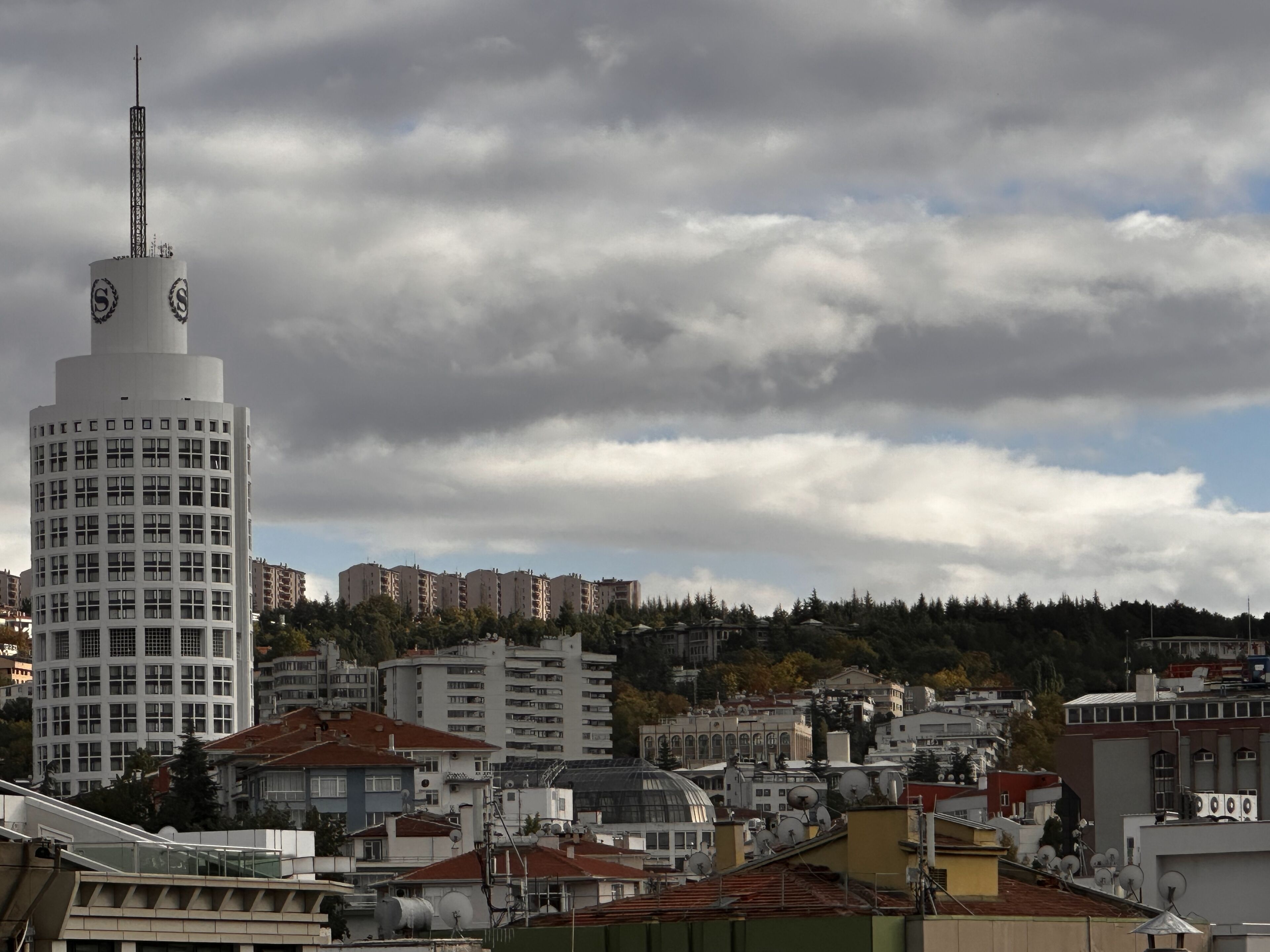 Paisagem urbana com uma torre branca imponente, que possui um relógio e uma antena, rodeada por edifícios residenciais e um cenário montanhoso.