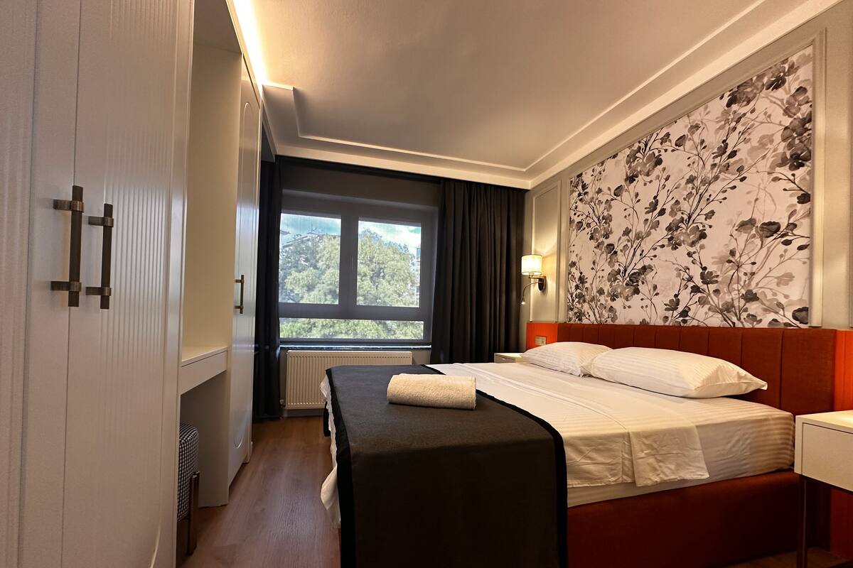 A hotel room with a bed, a bench, a window with a view, a radiator, and a wall with a floral pattern.