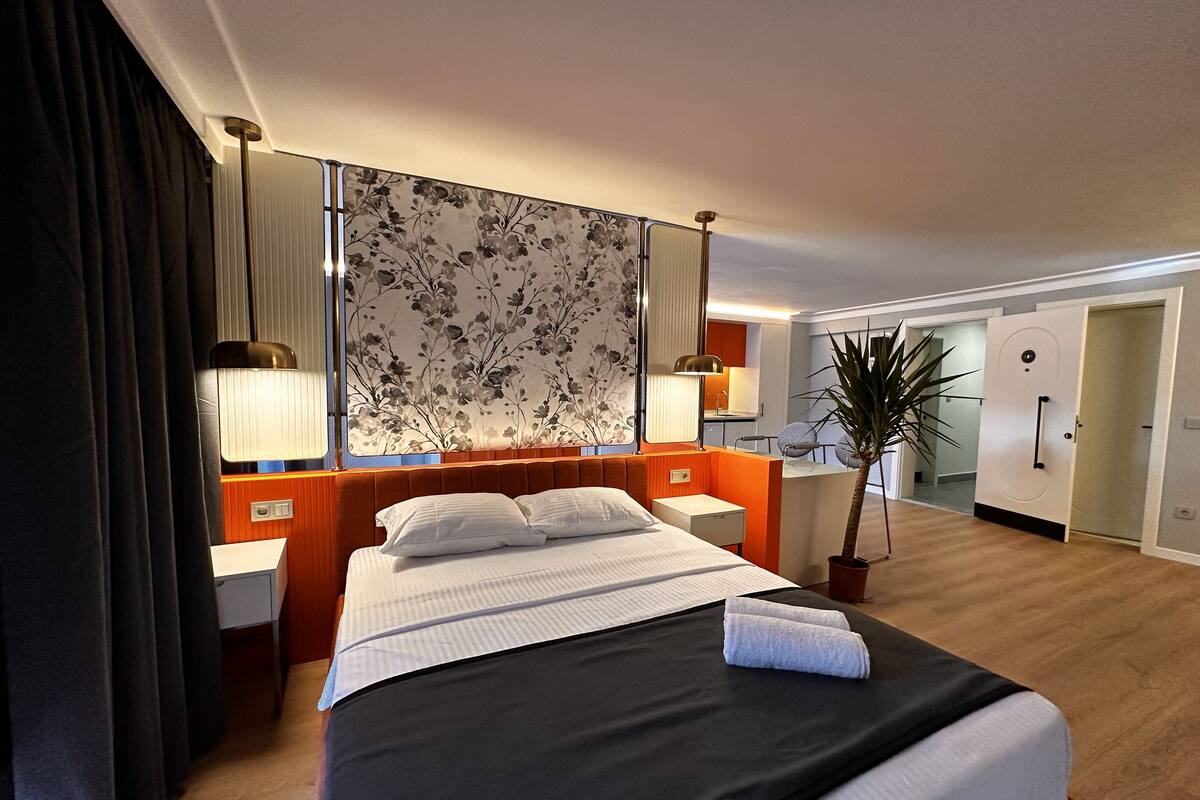 A hotel room with a bed, bedside tables, a nightstand, a lamp, a mirror, a potted plant, and a view of the bathroom.