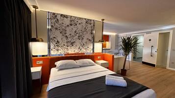 A hotel room with a bed, bedside tables, a nightstand, a lamp, a mirror, a potted plant, and a view of the bathroom.