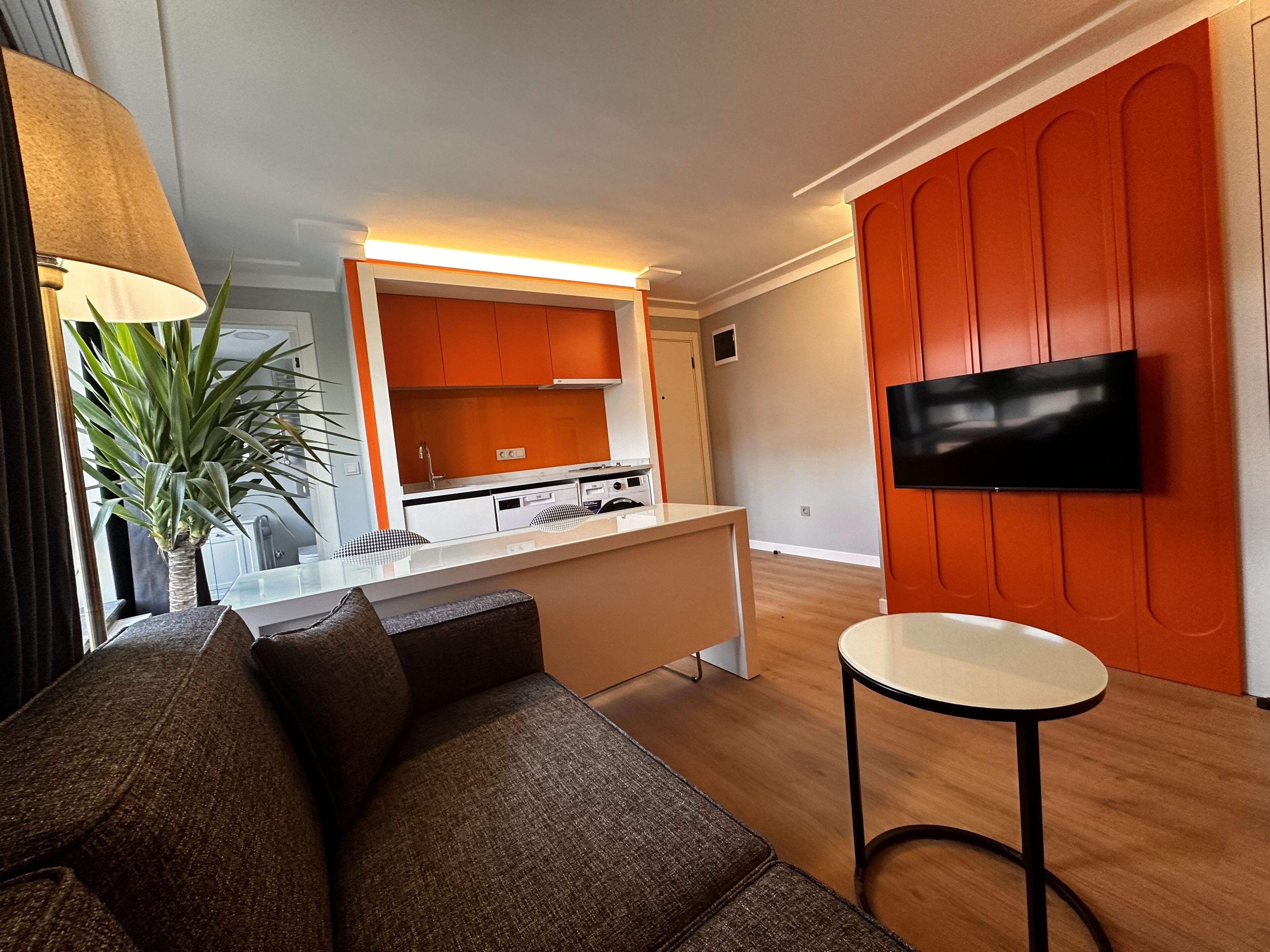 A modern living room with a sofa, a small round table, and a kitchen area with orange cabinets and a TV mounted on the wall.