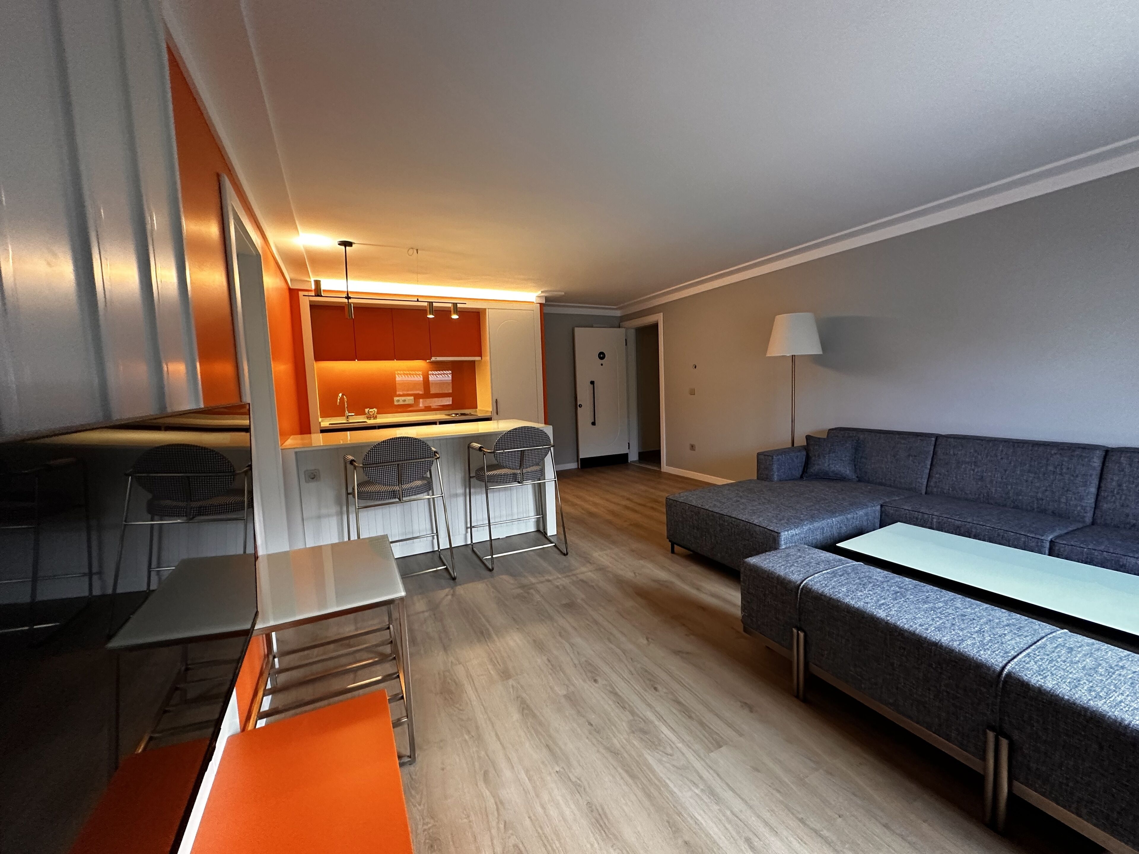 A modern living room with a grey sofa, a dining table with chairs, and a kitchen area with orange cabinets and a white countertop.