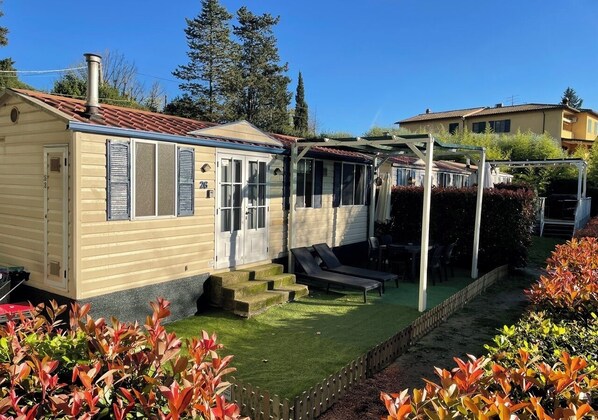 A mobile home with a small garden, a patio with a table and chairs, and a covered area with a roof. - Greener Camping Holiday Travel Genius at Toscana Holiday Village (San Romano)