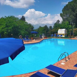 Piscina con sillas de descanso azules y sombrillas, rodeada de árboles y una zona cubierta.