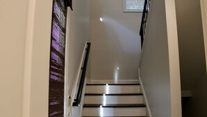 Un escalier aux murs blancs et aux marches sombres, menant à une pièce dotée d’une fenêtre et de stores.