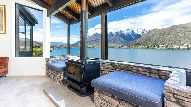 Ein gemütliches Zimmer mit Kamin aus Stein, großen Fenstern und Blick auf Berge und See.