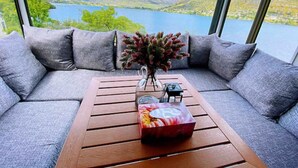 Ein modernes Zimmer mit großem Fenster mit Blick auf einen See und Berge. Vor einem Holztisch steht ein graues Sofa, auf dem Tisch steht eine Vase mit Blumen.
