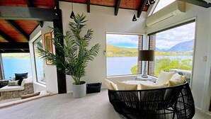 Ein Zimmer mit großem Fenster, einem schwarzen Gewebesessel, einer Topfpflanze und Blick auf einen See und Berge.