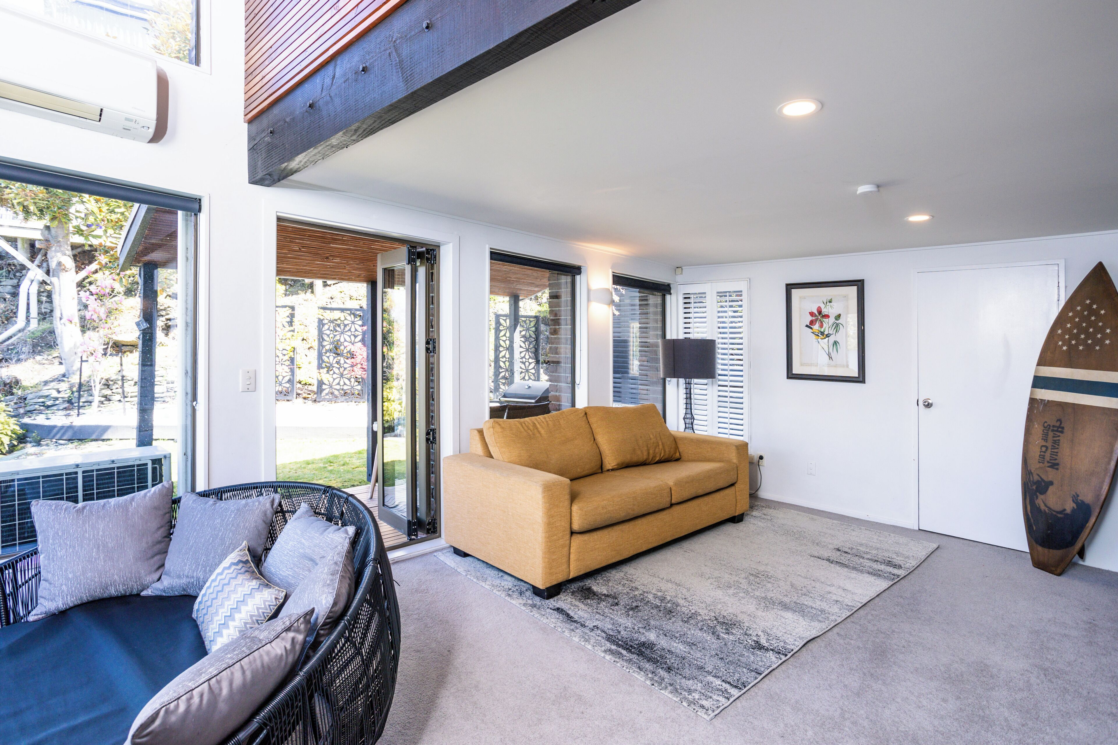 A modern living room with a sofa, a wicker chair, and a surfboard against the wall.