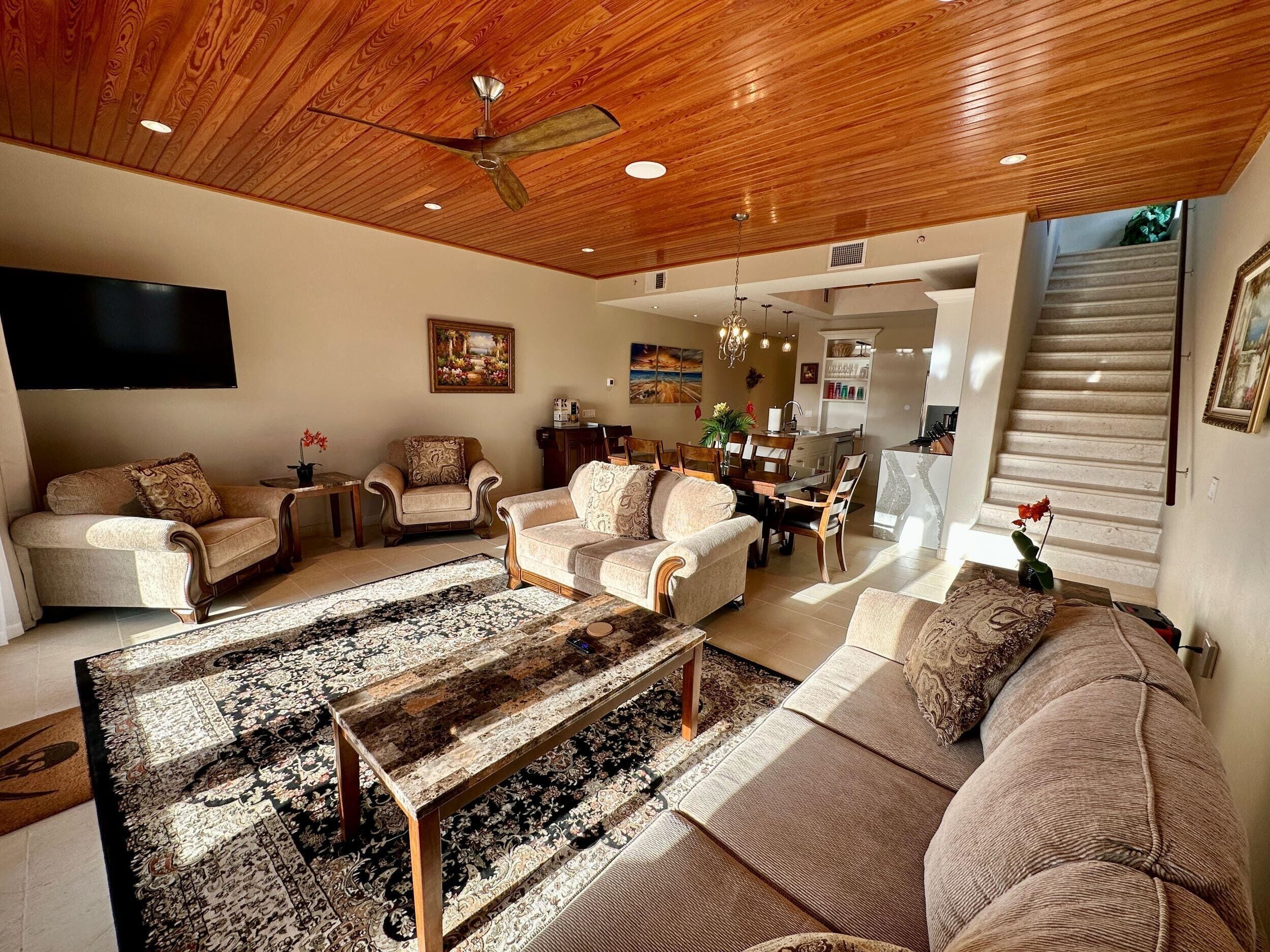 A spacious living room with a wooden ceiling, a large flat-screen TV, and a dining area with a kitchen in the background.