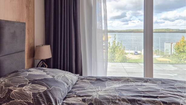 A bedroom with a large window overlooking a lake, a bed with a patterned bedspread, and a lamp on a bedside table. - Stunning apartment in Insko with sauna (Insko)