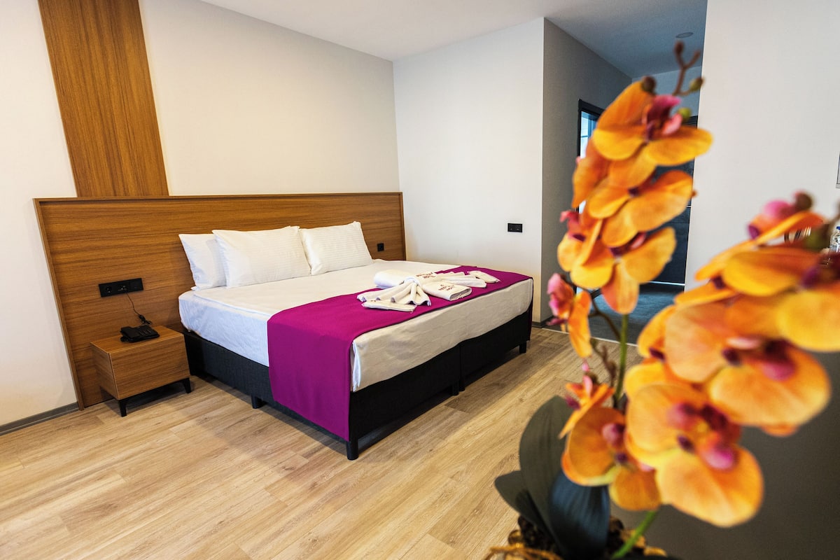 A hotel room with a wooden headboard, a bed with white linens and a purple blanket, a nightstand with a phone, and a wooden floor.